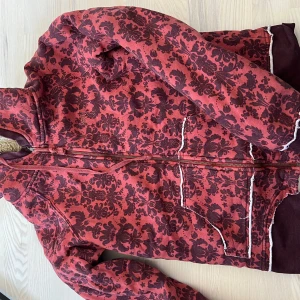 Röd mönstrad fleecejacka från Roxy - Säljer en röd fleecejacka från Roxy med vinrött blommigt mönster och mysigt teddyfoder. Jackan har dragkedja framtill, två fickor och vita detaljer längs kanterna. Perfekt för kyliga dagar!