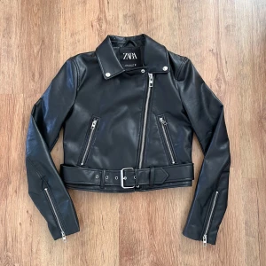 Snygg svart skinnjacka från Zara med bälte - Säljer en svart skinnjacka (fejk skinn) från Zara i bikerstil. Jackan har silvriga dragkedjor, bälte i midjan och klassisk krage. Croppad modell med långa ärmar och detaljerade sömmar. Perfekt för en edgy look. Jackan har ett litet hål. Om du är intresserad så kan jag skicka bild på det. 