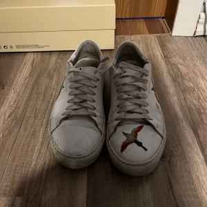 Vita sneakers med fågelmotiv från Axel Arigato - Säljer ett par vita sneakers från Axel Arigato med färgglada broderade fåglar på sidorna och tån. Skorna har snörning och platt sula, samt klassisk rund tå. Perfekta för dig som gillar stilrena men ändå unika detaljer.
