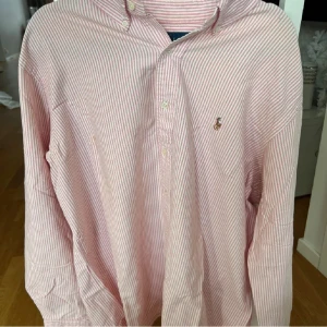 Röd- och vitrandig skjorta från Polo Ralph Lauren - Klassisk långärmad skjorta från Polo Ralph Lauren i röd- och vitrandigt mönster. Skjortan har knappar framtill och en broderad logga på bröstet. Perfekt för en stilren och avslappnad look.