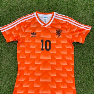 Nederländerna Gullit Fotbollströja EM 198 - Säljer en ikonisk orange Holland fotbollströja från Adidas med nummer 10 och namnet Gullit på ryggen. Tröjan har korta ärmar, vit krage och klassiska vita ränder på axlarna. Geometriskt mönster och svart tryck framtill och baktill.