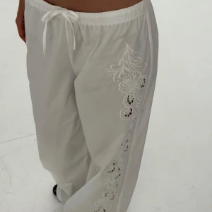 Vita broderade byxor från NA-KD - Säljer ett par vita byxor från NA-KD likt linnebyxor och vackra broderade blommor längs sidorna. Byxorna har resår och snörning i midjan för extra komfort. Perfekta för en somrig och avslappnad stil. Se sista bilden för lite lite smuts längst ned men går bort i tvätten. De är helt nya med lappen kvar! Slutsålda från chloe Schutermans kollektion. Storlek 38 passar S och M