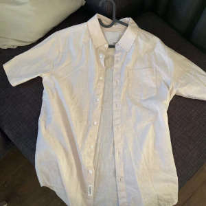 Beige kortärmad skjorta från H&M - Fräsch vit skjorta från H&M med korta ärmar och klassisk krage. Skjortan har knappar framtill och en bröstficka. Perfekt för varma dagar och enkel att matcha med olika outfits.