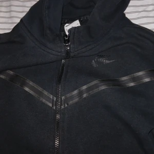 Svart Nike tech fleec - Säljer en svart hoodiejacka från Nike med dragkedja framtill och diskret Nike-logga på bröstet. Jackan har huva, långa ärmar och svarta detaljer längs bröstet och på ärmen. Perfekt för en sportig och avslappnad stil.