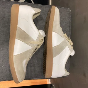 Maison Margiela skor - Säljer ett par stilrena sneakers i vitt och beige med snygga mockadetaljer och klassisk gummisula. Skorna har snörning och en platt sula, perfekt för en clean och avslappnad look. Passar till det mesta och är riktigt bekväma. Knappt använda och i toppen skick. Obs inte org1nal.