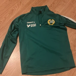 Grön långärmad träningströja från Craft Hammarby - Grön långärmad träningströja från Craft med Hammarby IF-emblem på bröstet och vit Intersport-logga. Tröjan har halv dragkedja framtill och vita ränder längs axlarna. Perfekt för fotbollsträning eller sportiga aktiviteter.