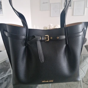 Svart handväska från Michael Kors - Stilren svart handväska från Michael Kors i äkta skinn. Väskan har guldiga detaljer, bälte med spänne framtill och innerfack med dragkedja. Perfekt för dig som vill ha en rymlig och klassisk accessoar. Nypris: 4400kr