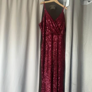 Vinröd paljettklänning med slits - Säljer en vinröd långklänning täckt av paljetter med smala axelband och omlott-v-ringning. Klänningen har en hög slits på sidan och är perfekt för festliga tillfällen. Materialet är glittrigt och skimrande, vilket ger en lyxig känsla. 