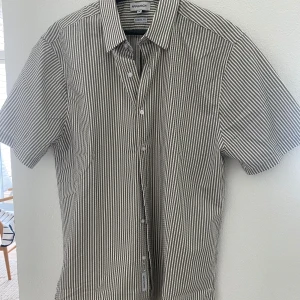 Randig kortärmad skjorta från Approach - Säljer en snygg kortärmad skjorta från Approach i storlek M. Skjortan har smala vita och gröna ränder, klassisk krage och knäppning framtill. Perfekt för en avslappnad stil.