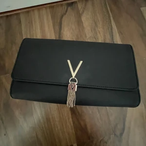 Svart valentino väska - Säljer en stilren svart kuvertväska från Valentino med guldfärgad kedja och dekorativt V-spänne framtill. Väskan har en elegant tofsdetalj och magnet lås!! Pris går att diskutera och bara använd 1 gång!!
