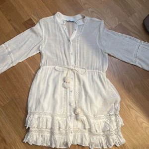 Vit kortklänning med volang och knytband - Söt vit kortklänning med långa ärmar, volangdetaljer och knytband i midjan med tofsar. Klänningen har dekorativa knappar framtill och spetsdetaljer längs volangerna. Perfekt för sommarens alla tillfällen.