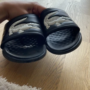 Svarta sandaler från Lacoste - Svarta sandaler från Lacoste med den klassiska vita krokodilloggan på ovansidan. Skorna har öppen tå och en räfflad sula för extra komfort. Perfekta för sommaren och enkla att ta på sig.Jävligt sköna för dem är extra mjuka.Priset går å diskutera