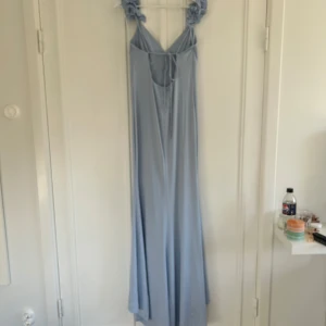 Ljusblå långklänning med volangaxel och knyt - Säljer en ljusblå långklänning med volangdetaljer på axelbanden och knyt framtill. Klänningen har en djup v-ringning och är ärmlös, perfekt för sommarens tillfällen. Materialet känns mjukt och följsamt.