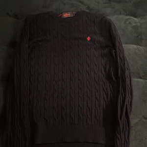 Svart kabelstickad tröja från Morris - Svart kabelstickad tröja från Morris i 100% merinoull. Tröjan har rund halsringning, lång ärm och en röd broderad logga på bröstet. Perfekt för lager-på-lager och stilrena outfits.