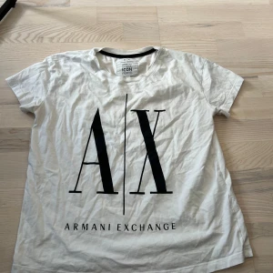 Vit t-shirt från Armani Exchange - Säljer en vit t-shirt från Armani Exchange med stor svart logga 'A|X' framtill. T-shirten har rund halsringning och korta ärmar. Perfekt för en stilren och trendig look.
