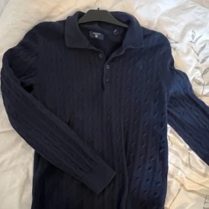 Gant stickad pikétröja - Säljer en mörkblå stickad pikétröja från Gant med klassisk krage och knappar framtill. Tröjan har lång ärm och ett diskret kabelstickat mönster som ger en stilren look. Perfekt för dig som gillar tidlös stil.