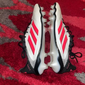Adidas Copa fotbollsskor  - Säljer ett par Adidas Copa fotbollsskor i vitt med svarta och röda detaljer. Skorna har klassisk snörning, rund tå och platt sula med dobbar för bra grepp på planen. Perfekta för dig som vill sticka ut på fotbollsplanen! Skorna är i storlek 46 men dem har blivit mindre och är ungefär 44-45