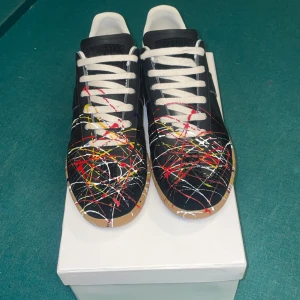 Maison Margiela sneakers med färgstänk - Maison Margiela sneakers i svart skinn med vita snören och unika färgstänk i rött, gult och vitt över hela skon. Klassisk rund tå och platt sula. Kommer med originalbox och dustbags. Perfekt för dig som vill sticka ut med din stil.