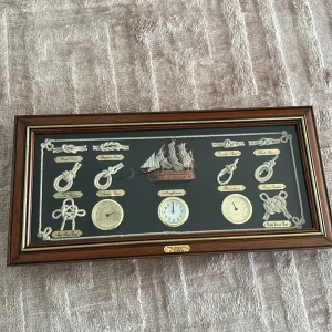 Maritim väggklocka med skepp och knopar - Unik väggklocka från Mega Gastronomi med maritimt tema. Klockan är inramad i mörkbrunt trä och har detaljer som en modell av ett segelfartyg och olika knopar i vitt rep. Urtavlan är vit med guldfärgad ram och det finns även barometer och termometer.