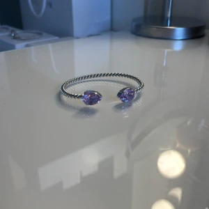 Caroline Svedbom armband - Snyggt armband i silverfärgad metall med tvinnad design och två stora, droppformade lila stenar i ändarna från Caroline Svedbom. Perfekt statement-piece för dig som gillar att sticka ut med accessoarerna. 💜💜