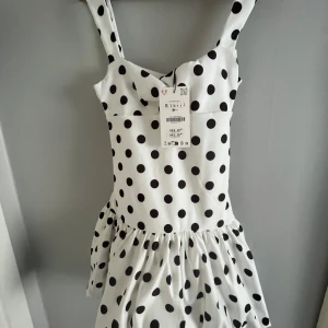 Vit prickig kortklänning S - Supersöt vit kortklänning med svarta prickar och breda axelband. Klänningen har en figurnära överdel och voluminös kjol med lätt rynk. Perfekt för dig som gillar retrovibbar och lekfull stil.