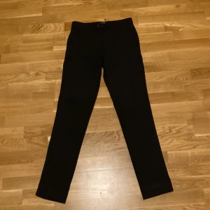 Svarta slim kostymbyxor Jack & Jones - Svarta kostymbyxor från Jack & Jones i modellen Slim/Marco. Byxorna har smal passform, klassisk design med bälteshällor och knappstängning. Perfekta för en clean och stilren look. Storlek 28/32.