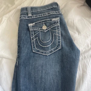 True Religion blå jeans med kontrastsömmar - Säljer ett par blå jeans från True Religion med ikoniska vita kontrastsömmar och snygga detaljer på bakfickan. Saknar en knapp på ena bakfickan, annars i bra skick!