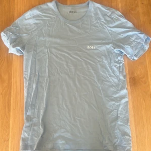 Ljusblå t-shirt från Hugo BOSS - Snygg ljusblå t-shirt från Hugo BOSS med klassisk rund hals och diskret BOSS-logga broderad på bröstet. T-shirten är i mjuk bomull och har en clean, enkel look som passar perfekt till jeans eller shorts.