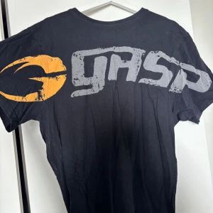 Svart GASP t-shirt med tryck - Svart t-shirt från GASP med stort grått logotryck över bröstet och en gul detalj på ärmen. Klassisk passform och rund halsringning. Perfekt för dig som gillar sportiga och streetinspirerade plagg.