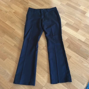 Svarta lågmidjade kostymbyxor bootcut - Snygga svarta kostymbyxor med bootcut passform och pressveck. Byxorna har klassisk passform med hällor för bälte och stängs med dragkedja och knapp. Perfekta för en clean och stilren look. Byxorna är väldigt lågmidjade och är i storlek 42 tall. Pris kan diskuteras.