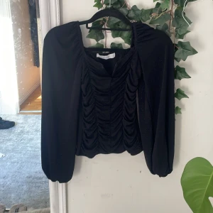 Svart glittrig blus från Sisters Point - Supersnygg svart blus från Sisters Point med glittriga detaljer och rynk framtill. Blusen har långa puffiga ärmar och en lätt v-ringad halsringning. Perfekt för dig som vill sticka ut lite extra med en cool och trendig look.