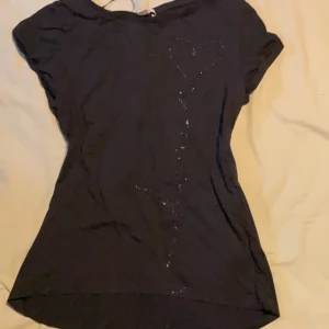 Svart topp med hjärtdetalj från Esprit - Svart topp från Esprit i mjuk bomull med glittrig hjärtdetalj framtill. Modellen har korta ärmar och en något lös passform. Perfekt för dig som gillar enkla plagg med en liten twist.