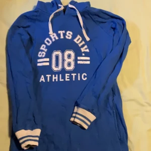 Lång blå hoodie med tryck och huva XS - Blå hoodie i sportig stil med vitt tryck 'SPORTS DIV. 08 ATHLETIC' på bröstet. Tröjan har huva med vita snören och ribbade muddar med blå-vita ränder. Perfekt för en chill och avslappnad look.