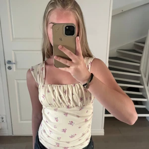 Blommig ljusgul linnetopp med rynk - Supersöt ljusgul linnetopp med små rosa blommor och rynkade detaljer vid bysten. Toppen har breda axelband och fyrkantig ringning både fram och bak. Perfekt för en somrig look och passar till både jeans och kjol.