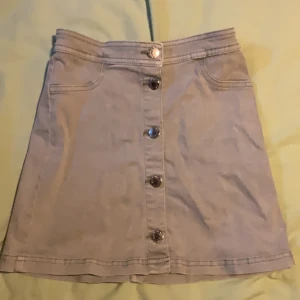 Beige kortkjol med knappar från H&M - Snygg beige kortkjol från H&M med raka linjer och fem coola knappar framtill. Kjolen har fickor fram och är gjord i ett mjukt bomullstyg som känns skönt mot huden. Perfekt för dig som gillar en enkel men trendig stil.