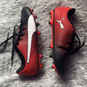 Röda och svarta Puma fotbollsskor - Säljer ett par snygga Puma fotbollsskor i storlek 38.5. Skorna har ett rött och svart sicksackmönster, vit Puma-logga på sidan och svarta snören. Yttersulan är röd med svarta dobbar, perfekt för gräsplan. Ovandelen är i syntetmaterial för bra passform.