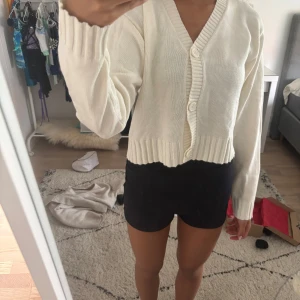 Beige Vit kofta från H&M med v-ringning - Beige vit kofta från H&M med v-ringning och stora knappar framtill. Modellen är croppad med bred ribbad mudd i ärmslut och nederkant. Långärmad och i mjuk stickad bomull, perfekt för lager-på-lager under höst och vår.