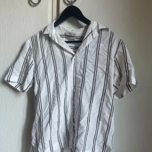 Vit randig skjorta från MNG Man - Vit skjorta från MNG Man med svarta och grå lodräta ränder. Regular fit-modell med korta ärmar och klassisk krage. Skjortan har knäppning framtill och är tillverkad i ett lätt bomullstyg, perfekt för en clean och stilren look. Aldrig använt skjortan  Pris kan diskuteras 