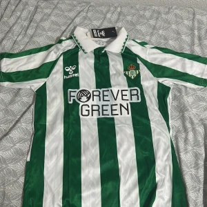 Real Betis Isco 22 Hummel fotbollströja - Snygg Real Betis fotbollströja från Hummel i storlek S men passar M, med gröna och vita vertikala ränder och tryck 'FOREVER GREEN' på bröstet. Isco och nummer 22 på ryggen. Klassisk krage och klubbmärke på bröstet. Perfekt för dig som älskar fotboll.