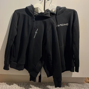 Sail racing 2x huv tröjor - Två svarta Sail Racing-plagg: en hoodie med stor logga framtill och känguruficka samt en ziphoodie med huva och diskret logga på bröstet. Båda har en clean look och är perfekta för dig som gillar sportig och stilren streetwear.