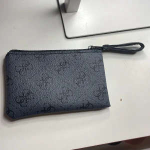 Guess plånbok - Snygg svart guess plånbok i skinnimitation med diskret mönster och dragkedja upptill. Perfekt att ha runt handleden tack vare den smidiga remmen. Enkel och stilren design som passar till många outfits. Köpt utomlands 