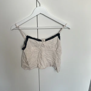 Beige linnetopp med spets från SHEIN - Söt beige linnetopp från SHEIN i storlek XS. Toppen har tunna axelband och är dekorerad med svart spets längs urringningen. Knappar framtill ger en extra detalj. Perfekt för en chill sommarlook.