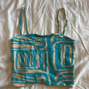 Turkos och beige linne med mönster - Säljer en snygg turkos linne-topp med tunna axelband och beige abstrakt mönster. Toppen är croppad och har raka linjer, perfekt för en cool och avslappnad stil. Materialet känns mjukt och lätt, perfekt för varma dagar.