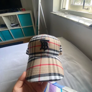 Rutig Burberry keps med TB-logo - Snygg keps från Burberry i klassiskt rutigt mönster med beige, svart, vitt och röda linjer. Framtill sitter en broderad svart TB-logo. Justerbar rem baktill för perfekt passform. Perfekt accessoar för att lyfta din stil.
