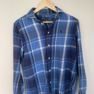 Blå rutig polo - Snygg blå rutig skjorta med långa ärmar och klassisk krage. Skjortan har knappar framtill och är i ett mjukt material som känns skönt mot huden. Perfekt för dig som gillar en avslappnad men ändå stilren look. Inga defekter