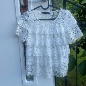 Vit spetsblus från Zara Basic - Superfin vit blus från Zara Basic i spets med korta ärmar och flera volanglager framtill. Blusen har rund halsringning och är genombruten i ett blommigt mönster. Perfekt för dig som gillar romantisk och luftig stil.