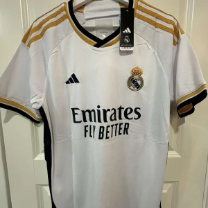 Real Madrid 25/26 Mbappé 9  - Snygg Real Madrid fotbollströja med Mbappé och nummer 9 på ryggen. Vit med guldiga och svarta detaljer, klassiska Adidas-ränder på axlarna och klubbmärke broderat på bröstet. Tillverkad i lätt och ventilerande material, perfekt för match eller träning.