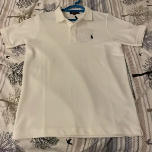 Vit pikétröja från Polo Ralph Lauren - Klassisk vit pikétröja från Polo Ralph Lauren med broderad svart logga på bröstet. Tröjan har korta ärmar, krage och knappar framtill. Tillverkad i bomull för en skön och luftig känsla. Perfekt för en clean och stilren look. Helt ny Ralph lauren piké som ja fick i present men den sitter tyvärr tight på mig så ja måste sälja vidare priset kan diskuteras! Skriv i dm om frågor och funderingar!