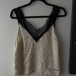 Beige linnetopp med svart spets - Snygg beige linnetopp från Stockh LM med breda axelband i svart spets och djup v-ringning både fram och bak. Toppens material är glansigt och känns lyxigt, perfekt för att lyfta vilken outfit som helst.