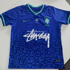 Blå Brasil x Stüssy x Nike fotbollströja - Snygg blå Brasil fotbollströja i samarbete med Stüssy och Nike⚡️. Tröjan har korta ärmar, gröna detaljer vid krage och ärmslut, samt ett leopardmönster nedtill. Materialet är glansigt och lätt, perfekt för fotboll eller streetwear. Stora Stüssy-loggan framtill och broderat Brasil-märke🦚. Pris är dikskuterbart. Hör av er vid frågor ❤️🌺✅